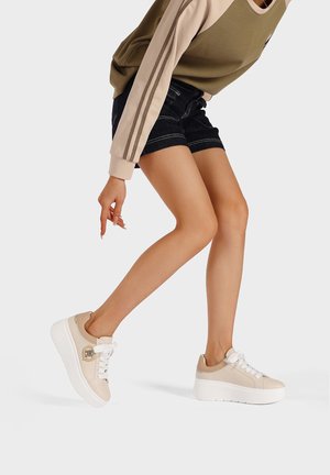 Sneakers beige con tomaia testurizzata, suola piatta bianca e dettagli decorativi. I lacci e il particolare del tallone a contrasto completano il design.
