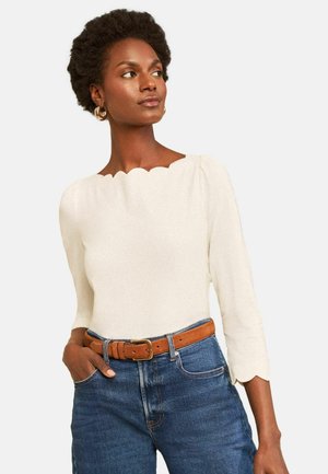 Woman with short curly hair wears ivory scalloped-edge top, blue jeans, and brown belt, standing with one hand in pocket, looking right.