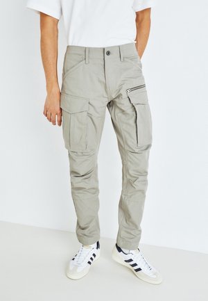 Pantalons cargo gris clair avec plusieurs poches, y compris une poche zippée sur la cuisse, fabriqués dans un tissu durable. Portés avec des baskets blanches.