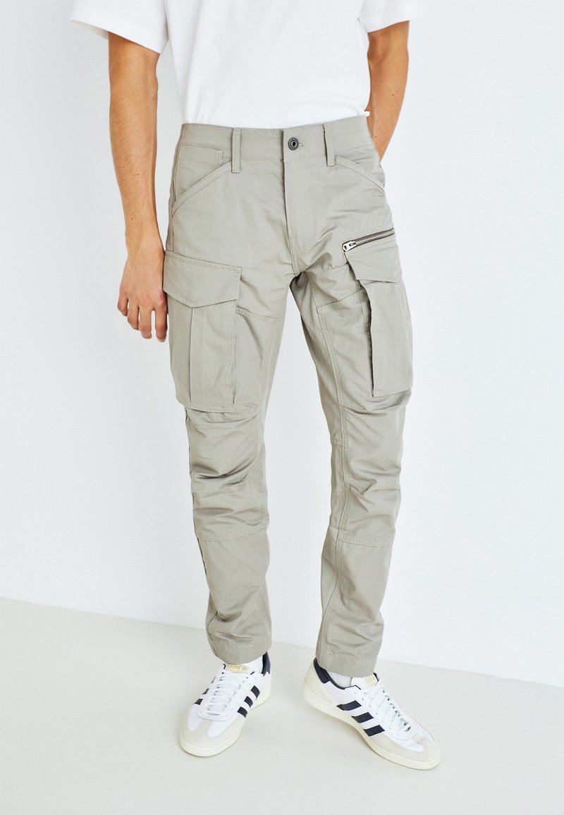 Světle šedé cargo kalhoty s několika kapsami, včetně zipové na stehně, vyrobené z odolného materiálu. Nosí se s bílými teniskami.