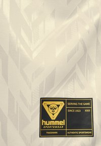 Witte stof met een getextureerd geometrisch patroon. Bevat een zwart-gouden label met "hummel" en merkdetails.