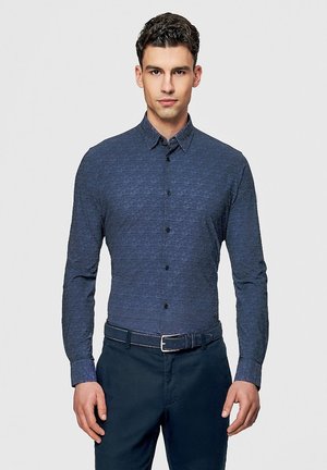 Mężczyzna zwrócony przodem, ubrany w dopasowaną niebieską koszulę zapinaną na guziki, włożoną do ciemnych spodni z pasującym paskiem, na tle jednolitego jasnego tła.