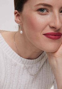 Srebrny naszyjnik z małymi perłami oraz dopasowanymi wiszącymi kolczykami z pojedynczą perłą. Modelka ma na sobie jasny, teksturowany biały sweter.