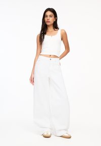 Weißes Tanktop mit rundem Ausschnitt, kombiniert mit weiten weißen Jeanshosen. Das Model trägt legere Schuhe und zeigt eine entspannte Passform.