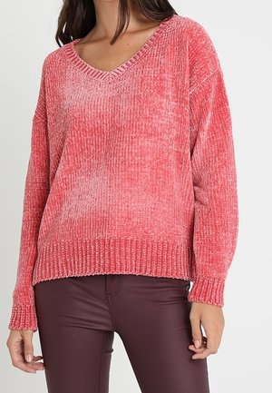 Woman wearing a soft, ribbed, pink V-neck sweater and burgundy fitted pants, standing against a plain light background.