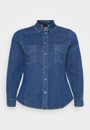 Camisa de ganga em azul escuro, com mangas longas, fecho de botão à frente, dois bolsos no peito e costuras visíveis.