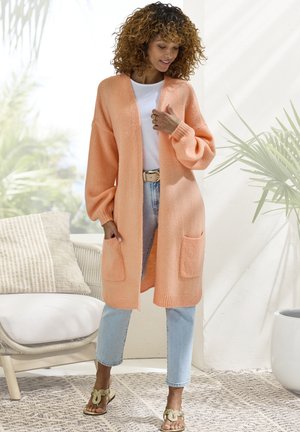 Peach knitted long cardigan with long sleeves, ribbed cuffs, and two front pockets, worn over a white shirt and light blue jeans.