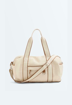 Geantă duffle din pânză bej cu tiv maro, mânere duble, bretea de umăr reglabilă, buzunar frontal cu fermoar și detaliu metalic în formă de D.