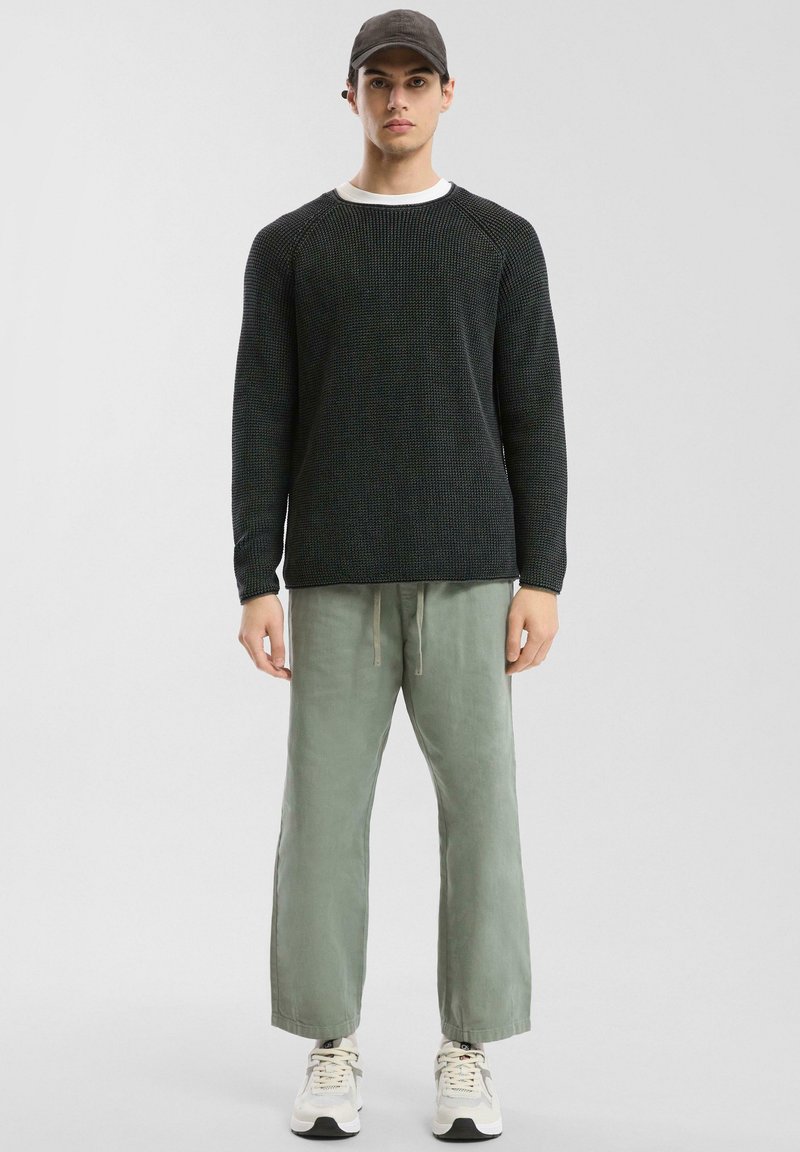 Pull en maille noir texturé, T-shirt gris clair en dessous, pantalon vert sauge avec cordon de serrage et baskets blanches avec accents noirs.
