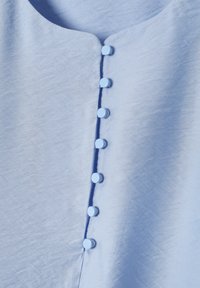 Blouse bleu clair avec un col arrondi, une texture lisse et six boutons ronds et brillants le long de la patte de boutonnage avant.