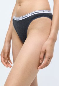 Szare bawełniane stringi z białym elastycznym pasem na biodra z logo "Calvin Klein". Gładka tekstura, minimalistyczny design, niski stan.