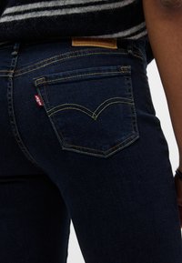 Κοντινή λήψη πίσω τσεπής σκούρου μπλε τζιν με κίτρινη ραφή και μικρή κόκκινη ετικέτα Levi's, φορεμένα με μαύρη και άσπρη ριγέ ζακέτα.