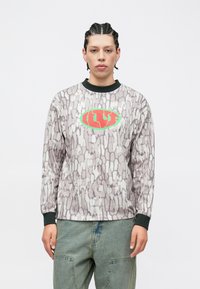 Mikina s dlhým rukávom s textúrovaným sivým vzorom, čiernym golierom a centrálnym červeno-zeleným logom. Kombinovaná so zelenými cargo nohavicami.