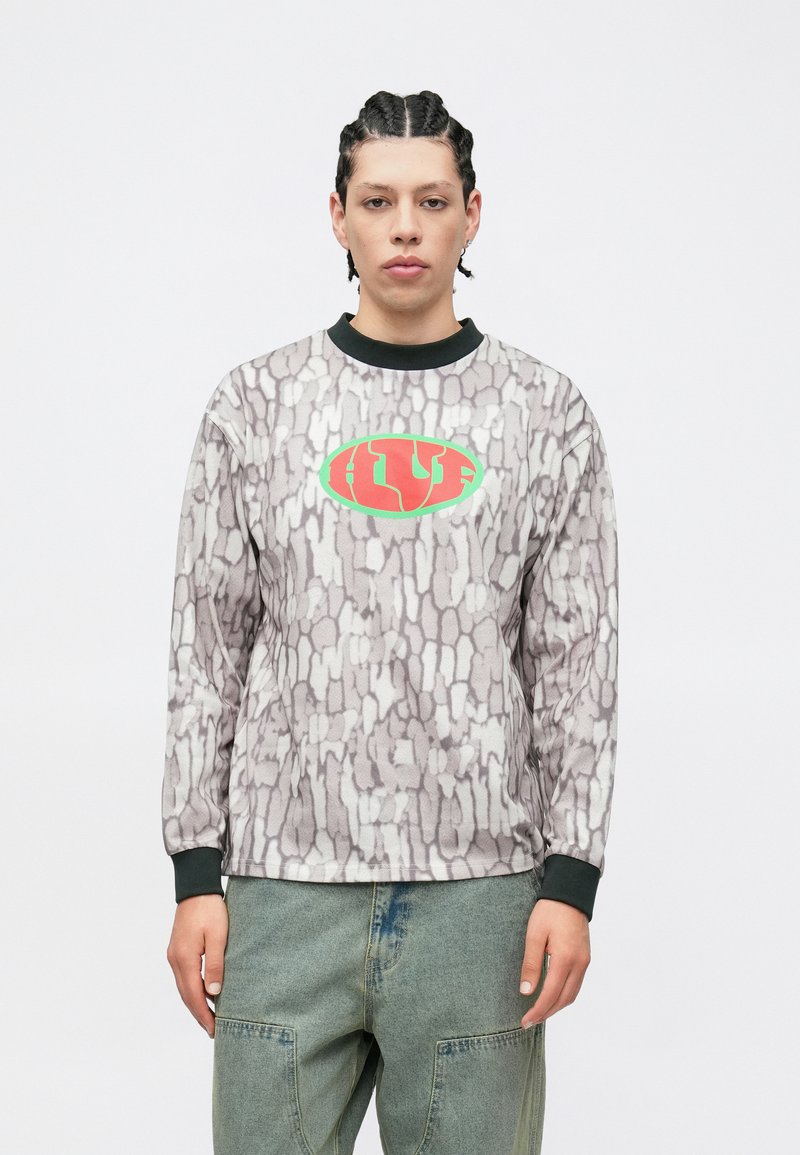 Mikina s dlhým rukávom s textúrovaným sivým vzorom, čiernym golierom a centrálnym červeno-zeleným logom. Kombinovaná so zelenými cargo nohavicami.