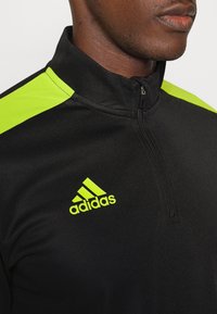 Svart polyesterjacka med dragkedja och gröna accenter på axlarna. Har Adidas-logotyp i ljusgrönt på bröstet. Mjuk textur.