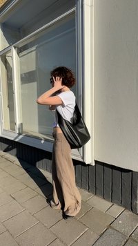 Črna usnjena hobo torba s ukrivljeno obliko in mehko teksturo. Model nosi belo majico in svetlo bež široke hlače, stoji na prostem.