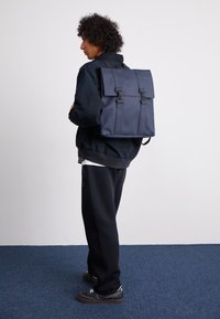 Sac à dos bleu marine fabriqué à partir de matériaux durables, doté d'une fermeture à rabat, de deux bretelles réglables et de quincaillerie en argent, avec un design minimaliste.