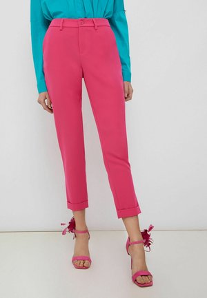 Femme portant un pantalon tailleur rose vif longueur cheville, associé à des sandales à talons hauts assorties ornées de plumes.