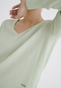 Pull en maille vert clair avec un col en V, des variations de texture avec des manches côtelées, et un petit détail de plaque en métal à l'ourlet.