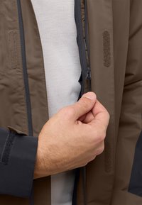 Bruin ritsjack met een gladde textuur. Bevat klittenbandaccenten op de mouwen en een close-up van een hand die de ritsgrijper vasthoudt.