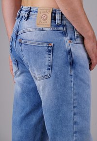 Hellblaue Jeans aus Denim mit einem verblassten Finish, ausgestattet mit zwei Gesäßtaschen, Kupferriegeln und einem braunen Lederschild am Bund.