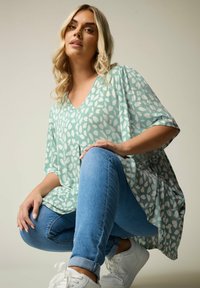 Mintgrön tunika med vitt bladmönster, V-ringning, lös passform, kombinerad med åtsittande blå jeans och vita sneakers. Slät tygstruktur.