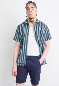 Chemise à manches courtes avec des rayures verticales bleues et vertes, associée à un short marine. La chemise dispose d'une poche poitrine et d'un col évasé.