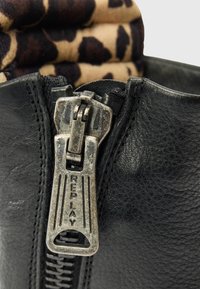 Κοντινό πλάνο από μια φθαρμένη ασημένια λαβή φερμουάρ με την επιγραφή "REPLAY" σε ένα μαύρο δερμάτινο ρούχο και ύφασμα με λεοπάρ σχέδιο στο εσωτερικό.