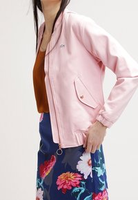 Giacca bomber rosa chiaro con polsini a costine, tasca laterale e chiusura con zip. Abbinate a una gonna con stampa floreale dai colori vivaci.