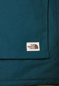 Tealfärgad tyg med en ficka och en vit rektangulär lapptagg med "THE NORTH FACE"-logotypen i brunt.