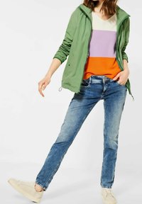 Ljusgrön lätt jacka över en flerfärgad randig tröja (vit, lavendel, orange) som matchas med blå jeans och ljusbeiga sneakers.