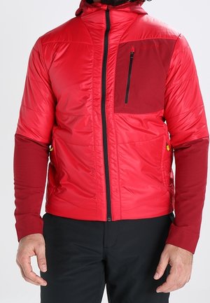 Rote isolierte Jacke mit dunkelroter Reißverschlusstasche auf der Brust und Ärmeln, getragen über einer schwarzen Hose, Hände entspannt an den Seiten.