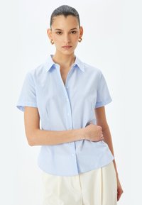 Lichtblauwe blouse met korte mouwen en witte verticale strepen, knoopsluiting aan de voorkant en een kraag. Gemaakt van een gladde, lichte stof.