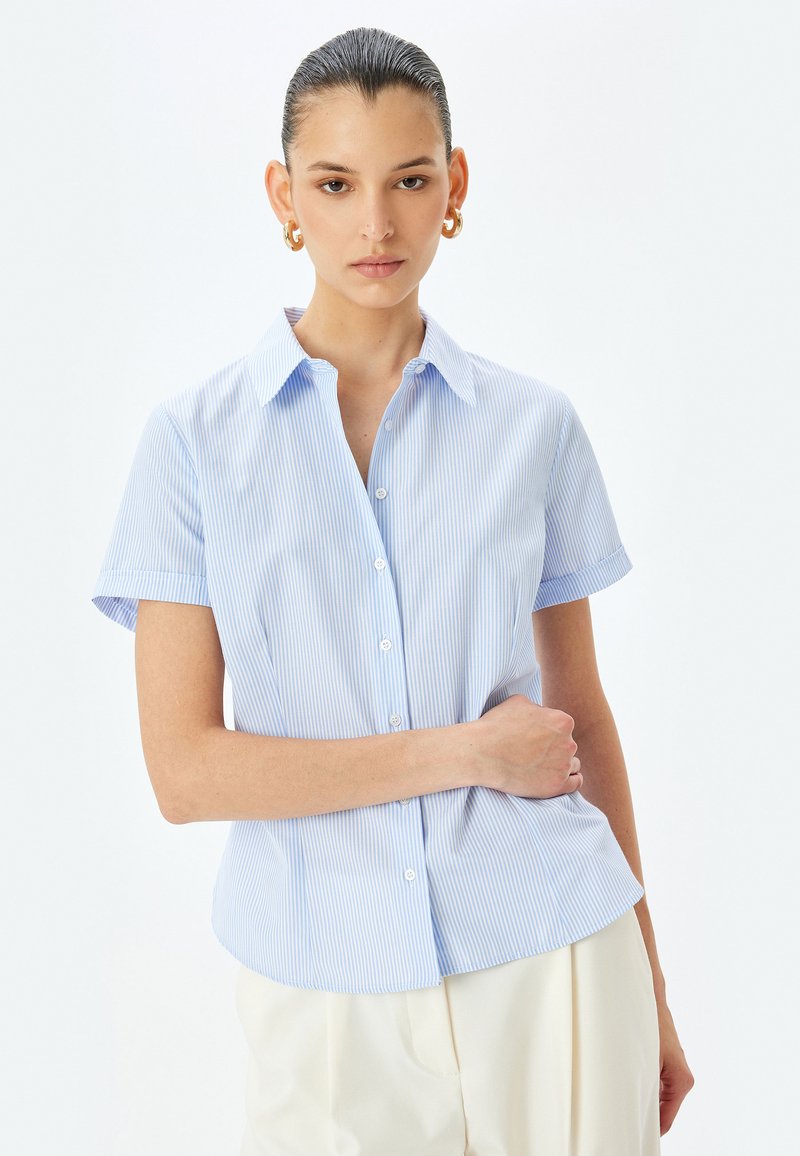 Blusa azzurro chiaro a maniche corte con righe verticali bianche, chiusura con bottoni frontale e colletto. Realizzata in un tessuto liscio e leggero.