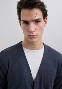 Cardigan gris charbon avec un décolleté en V profond et trois boutons, superposé à un t-shirt gris clair à col rond ; texture douce et design ajusté.