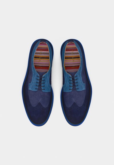 Paire de chaussures brogues en daim bleu pour hommes, à lacets, avec des semelles intérieures rayées multicolores, vues de dessus sur un fond blanc.