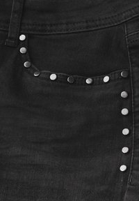 Tessuto di denim nero con una tasca frontale caratterizzata da borchie argentate lungo il bordo e accenti di borchie verticali lungo la cucitura laterale.