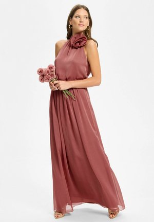 Donna con abito maxi senza maniche color rosa polvere con un grande dettaglio floreale su una spalla, che tiene dei crisantemi rosa, in piedi su sfondo bianco.
