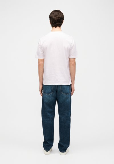 Άνδρας που στέκεται με την πλάτη, φοράει απλό λευκό κοντομάνικο πουκάμισο, μπλε τζιν και λευκά αθλητικά παπούτσια μπροστά σε λευκό φόντο.