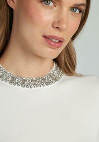 Maglione bianco con colletto di perline che presenta strass argento e trasparenti. Tessuto morbido, primo piano che rivela i dettagli del colletto e del viso.