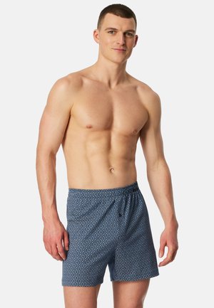 Muskuløs mand iført blå mønstrede boxershorts med elastisk taljebånd står foran en ensartet lys baggrund.