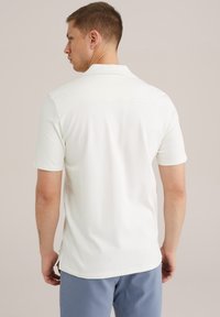 Polo alb cu mânecă scurtă, cu croială dreaptă, textură netedă și detaliu pe spate, asociat cu pantaloni ușor albaștri.