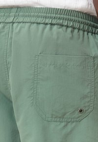 I pantaloni corti verdi con cintura elastica presentano una tasca posteriore con un design ottagonale e un eyelet metallico nell'angolo inferiore. Il tessuto è liscio.