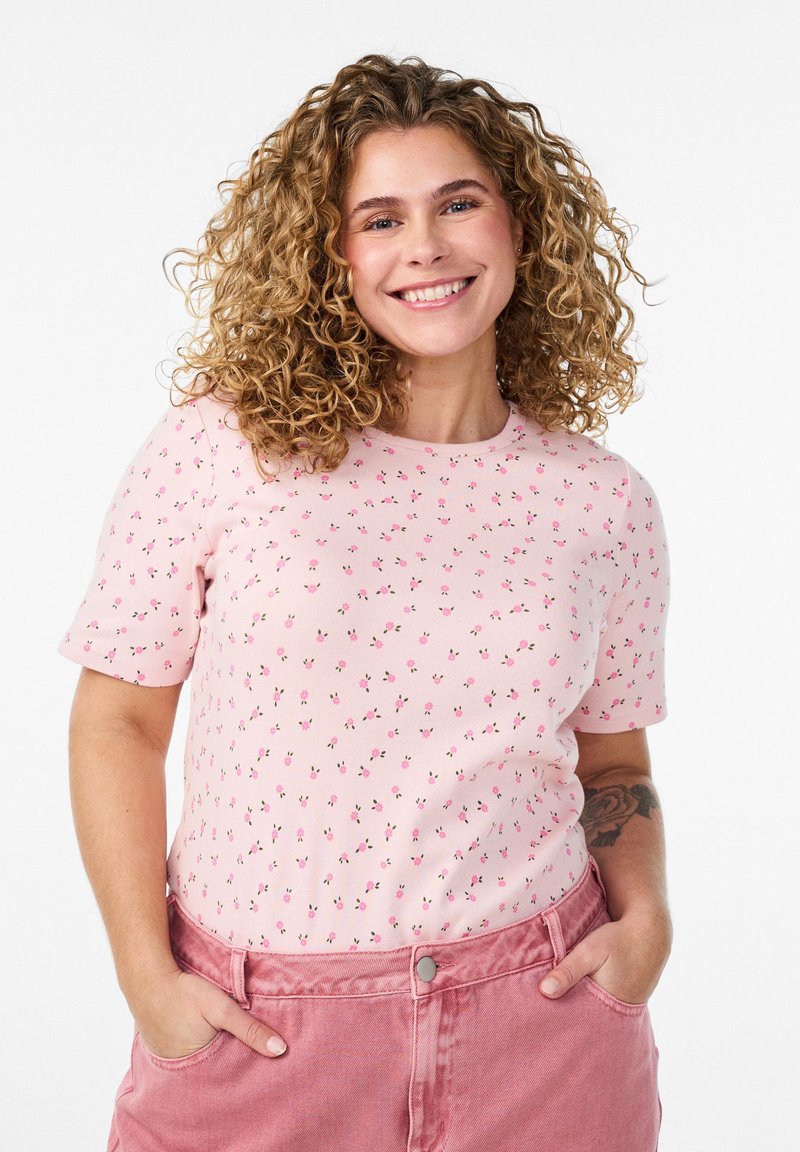 Smaidoša sieviete ar cirtainiem blondiem matiem, tērpusies rozā ziedu raksta īss piedurkņu krekliņā un rozā biksēs, rokas makos, balts fons.