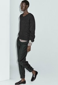 Kvinna som bär en svart texturerad tröja, grå skjorta under, åtsittande svarta byxor, svarta loafers och ett silverarmband, som står mot en vit vägg.