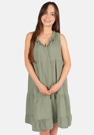Jeune femme souriante, portant une robe sans manches vert clair à volants avec un col froncé et des liens à pompons, mains délicatement jointes.