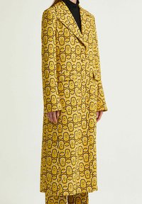 Cappotto giallo con motivi geometrici marroni e bianchi, caratterizzato da un colletto a risvolto, maniche lunghe e due tasche frontali.