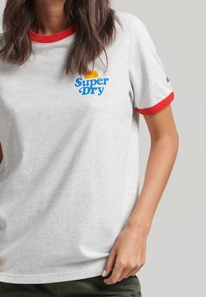 Kobieta nosząca białą koszulkę z czerwonym kołnierzykiem i mankietami, z małym logo "Super Dry" i grafiką słońca na piersi.