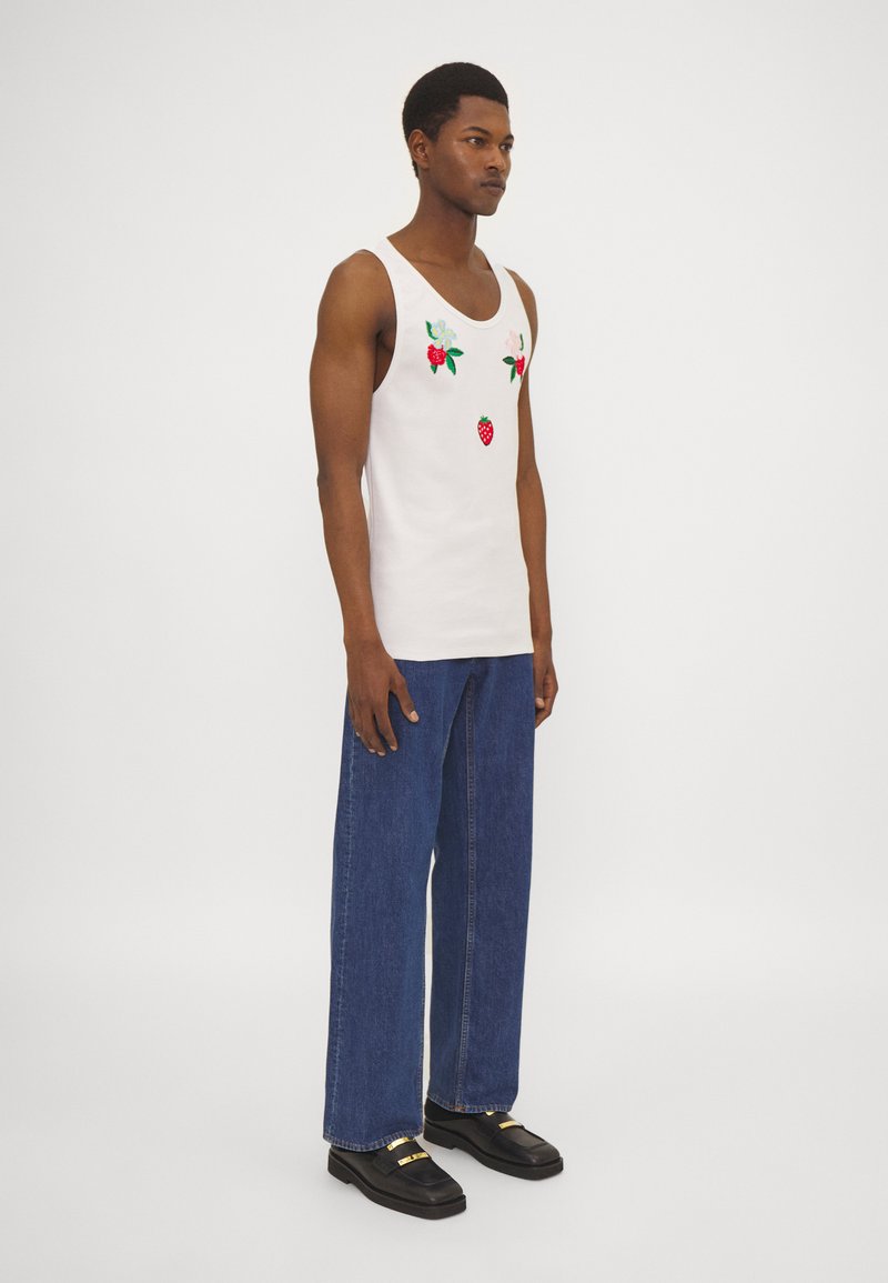 Top blanco de tirantes con bordado de fresas y flores, combinado con pantalones vaqueros de pierna ancha azules y zapatos negros.
