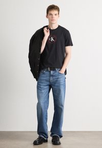 Czarny bawełniany T-shirt z czerwonym, okrągłym logo CK, w połączeniu z jasnoniebieskimi dżinsami i czarnymi butami, trzymający czarną kurtkę na ramieniu.